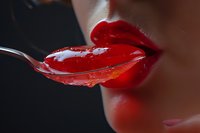 Close-up of woman's lips with bright red lipstick tasting sweet cherry jelly from a spoon against dark background.