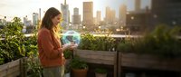 Young woman tending to urban rooftop garden with city skyline at sunset. Professional gardener watering plants in modern container garden for sustainable living.