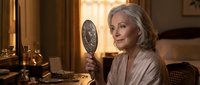 Elegant mature woman with silver hair looking in vintage hand mirror in warm-lit classic interior with antique furnishings and golden lighting.