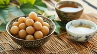 Traditional Asian sweet rice balls in decorative ceramic bowl with tea cups on bamboo mat, surrounded by green leaves for authentic presentation.
