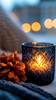 Glowing candle in blue textured glass holder with pine cone and cozy knit texture, creating warm atmospheric lighting against soft bokeh background.