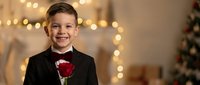 Young Caucasian boy in formal black tuxedo holding red rose with warm bokeh lights and Christmas tree creating elegant holiday portrait atmosphere.