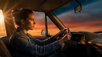 Young Caucasian female soldier in military uniform driving vehicle during golden hour sunset with dramatic sky creating cinematic atmosphere for patriotic content.