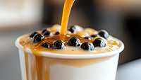 Caramel sauce pouring over creamy bubble tea topped with black tapioca pearls in white cup, close-up view showing rich golden syrup and glossy boba texture.
