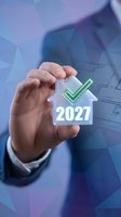 Businessman hand holding holographic house icon with green checkmark and 2027 year number against blue geometric background for real estate, property investment, and home approval concepts.