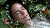 Young woman with closed eyes lying among green leaves and soap bubbles, expressing serenity and natural beauty in dreamy garden atmosphere.
