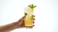 Hand holding refreshing yellow lemonade cocktail with mint leaves and ice cubes in tall glass against white background for summer beverage designs.