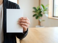 Person of Middle Eastern or South Asian descent holding blank white paper sheet in office environment with potted plant and wooden desk for mockup.
