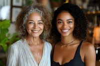 Joyful portrait of mature woman with gray curly hair and young African American woman with natural curls sharing genuine smiles in cozy home environment.