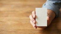 Close-up of a person's hand holding a blank white card or paper against a wooden surface, suitable for branding, business cards, or mockup presentations.