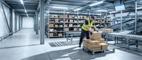 Male warehouse worker in safety vest organizing cardboard boxes on pallet in modern distribution center with metal shelving and industrial lighting.
