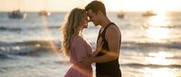 Young couple embracing intimately on beach at golden hour sunset with ocean waves and boats in background creating romantic atmosphere for wedding or engagement.
