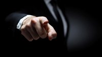 Businessman in formal suit pointing finger directly at viewer against dark background, conveying authority and leadership in corporate environment.