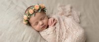 Peaceful newborn baby girl sleeping wrapped in soft pink lace blanket wearing delicate floral crown headband on textured cream backdrop for portrait photography.
