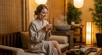 Young Caucasian woman in beige bathrobe relaxing at luxury spa with herbal tea, enjoying wellness treatment in serene bamboo interior with warm lighting.