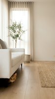 Curious ginger tabby cat hiding under a white sofa in a bright minimalist living room with natural light, jute rug, and indoor plant.