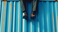 Businessman legs in formal suit and dress shoes sitting on blue striped container surface with orange accents in modern urban setting.