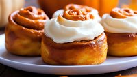 Freshly baked cinnamon rolls topped with smooth cream cheese frosting and caramel swirl on white plate, close-up view showing soft, fluffy texture and golden surface.
