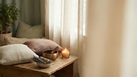Cozy bedroom interior with burning candle on wooden nightstand, soft pillows and warm natural lighting creating peaceful atmosphere for relaxation.