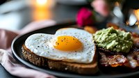 Heart-shaped fried egg on toasted bread with fresh guacamole and crispy bacon strips on dark plate, morning breakfast setting with soft bokeh lights.