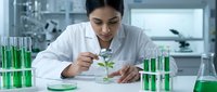 Female scientist examining green plant seedling with magnifying glass in modern laboratory surrounded by test tubes containing green liquid samples for botanical research.