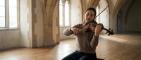 Young Asian woman violinist playing violin in historic stone architecture hall with arched windows and wooden floor for classical music performance.
