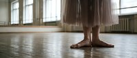 Young ballerina feet in ballet slippers standing on wooden dance studio floor with tulle skirt and natural window light creating artistic atmosphere.