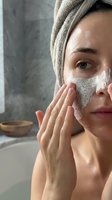 Young woman applying exfoliating face scrub mask in modern bathroom. Natural skincare routine, spa treatment at home for healthy glowing skin.