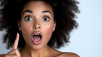 Young African American woman with natural curly afro hair expressing surprise with open mouth and raised finger against light background. Emotional portrait.