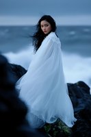 Young Asian woman in flowing white dress stands on coastal rocks against stormy sea, dark moody atmosphere with wind-swept hair creates mystical scene.