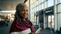 Confident African American businesswoman with dreadlocks holding tablet in modern airport terminal, smiling at camera while traveling for work.