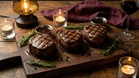 Grilled beef steaks with perfect grill marks on wooden cutting board, garnished with fresh rosemary herbs, surrounded by candlelight and red wine for elegant dining.