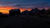 Abandoned wooden cabin silhouetted against dramatic orange and purple sunset sky with jagged mountain peaks in remote wilderness landscape at dusk.