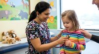 Female pediatric nurse in colorful scrubs examining young Caucasian girl patient with stethoscope in bright medical office with animal wall decorations.