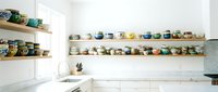 Modern minimalist kitchen interior with open wooden shelving displaying colorful ceramic bowls and dishes against white walls and countertops.