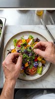 Chef plating colorful salad with microgreens and edible flowers on ceramic dish. Overhead view of fine dining food preparation with fresh vegetables and garnish.