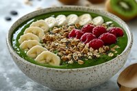 Green smoothie bowl topped with sliced banana, fresh raspberries, granola, and seeds in ceramic bowl. Healthy breakfast with superfood ingredients.