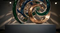 Modern abstract glass sculpture with copper metallic accents on white pedestal in gallery setting with dramatic lighting and shadows for contemporary art displays.