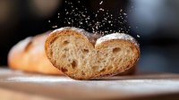 Fresh crusty baguette slice with flour dust falling in dramatic lighting against dark background, close-up view showing detailed bread texture and crumb structure.