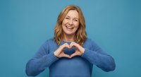 Happy blonde woman making heart shape with hands wearing blue sweater on blue background expressing love and positive emotions for lifestyle content.
