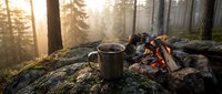 Metal camping mug sits on moss-covered rock beside crackling campfire in misty forest during golden hour sunrise creating peaceful outdoor adventure scene.