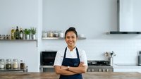 Asian female chef or restaurant owner wearing white shirt and dark apron, smiling with arms crossed in modern kitchen with white walls, shelving, and cooking equipment.