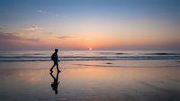 Silhouetted surfer carrying surfboard walking along wet beach sand at golden sunset, ocean waves in background, suitable for travel and lifestyle content.