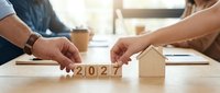 Two people arranging wooden blocks showing 2027 next to a small wooden house model on a desk, representing future real estate planning and investment goals.
