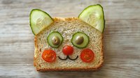 Playful cat face made from whole wheat bread slice decorated with cucumber ears, olive eyes and cherry tomato nose and smile. Fun food art for children.