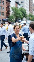 Middle-aged Caucasian female food vendor in blue apron serving customers on busy urban street with yellow taxis and office workers in background.