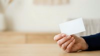 Mature man with medium skin tone holding blank white business card in hand at wooden table, suitable for corporate branding and professional networking concepts.
