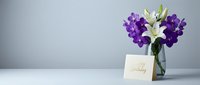 Purple orchids and white lily arrangement in glass vase with greeting card on clean white surface against soft gray background for sympathy condolences.