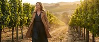 Young blonde woman walking through vineyard rows at golden hour sunset, wearing elegant coat, wine country lifestyle and agriculture tourism concept.