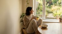 Young woman in beige loungewear sitting by window with coffee cup enjoying peaceful morning moment at home with natural light and plants.